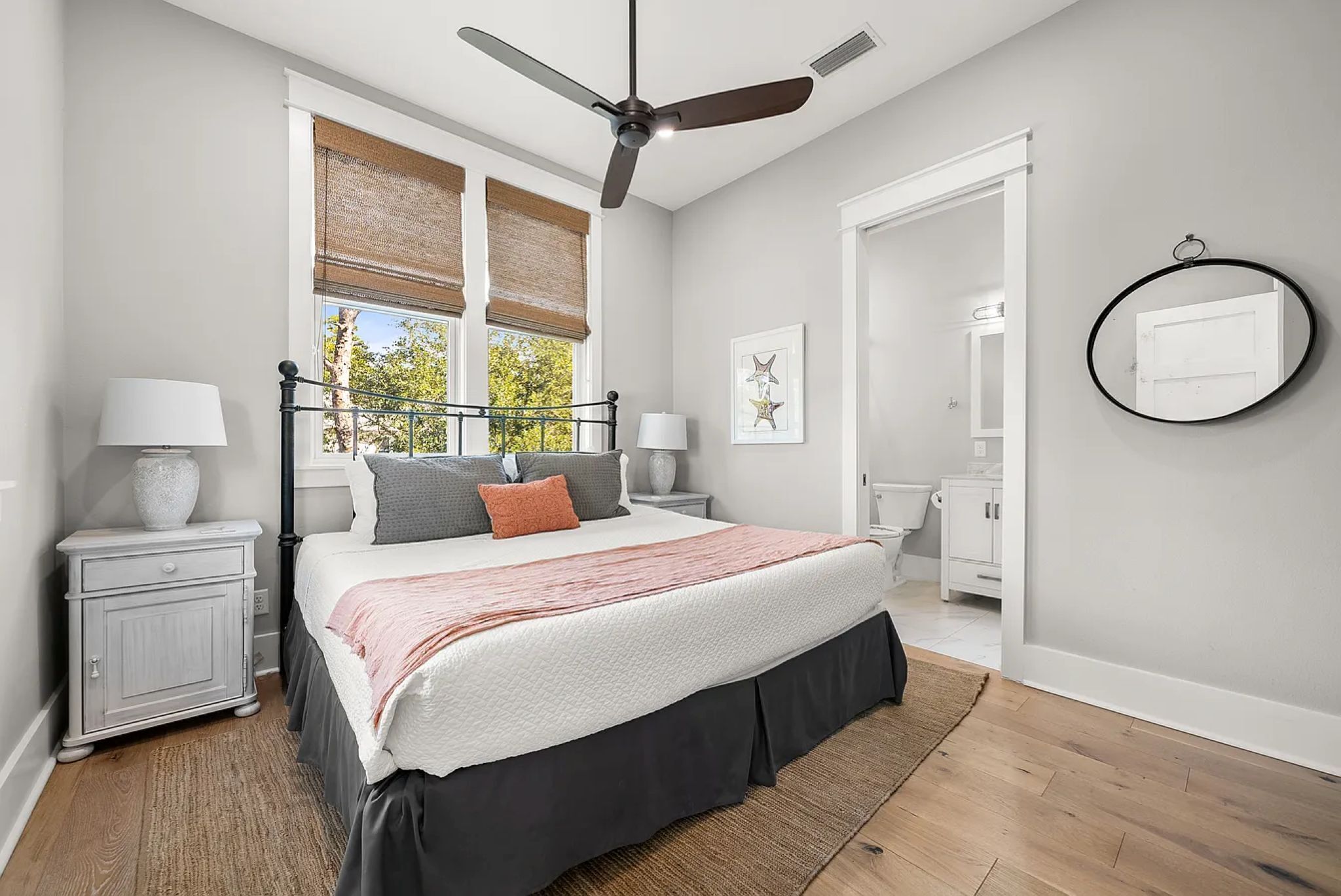 A bedroom with a bed, a ceiling fan, a window with blinds, a bedside table with a lamp, and a mirror on the wall.