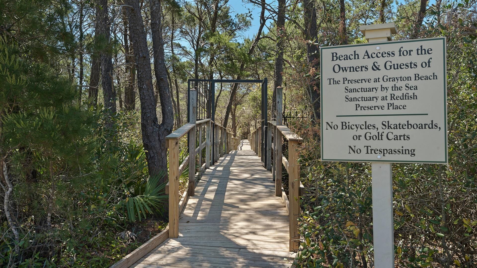 Ein hölzerner Steg führt zu einem Strand, umgeben von Bäumen. Ein Schild weist auf den Zugang zum Strand für Eigentümer und Gäste des Preserve at Grayson Beach Sanctuary by the Sea und des Sanctuary at Redfish Preserve Place hin.