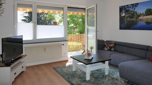 A living room with a TV on a stand, a sofa, a coffee table, and a large window with blinds. - New from 2022: beautiful 84 sqm vacation apartment in a top location with partial lake view (Langenargen)