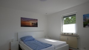 A bedroom with a single bed, a nightstand, a radiator, and a window with a view of greenery. - New from 2022: beautiful 84 sqm vacation apartment in a top location with partial lake view (Langenargen)