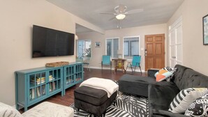 A living room with a TV, teal cabinet, ottoman, black sofa, patterned rug, and a ceiling fan. - Ben`s Beach Bungalow 3, Ocean View Mission Beach 1 (San Diego)