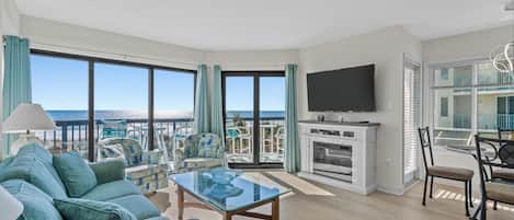 A spacious living room with a large TV, comfortable seating, a glass coffee table, and a view of the ocean.