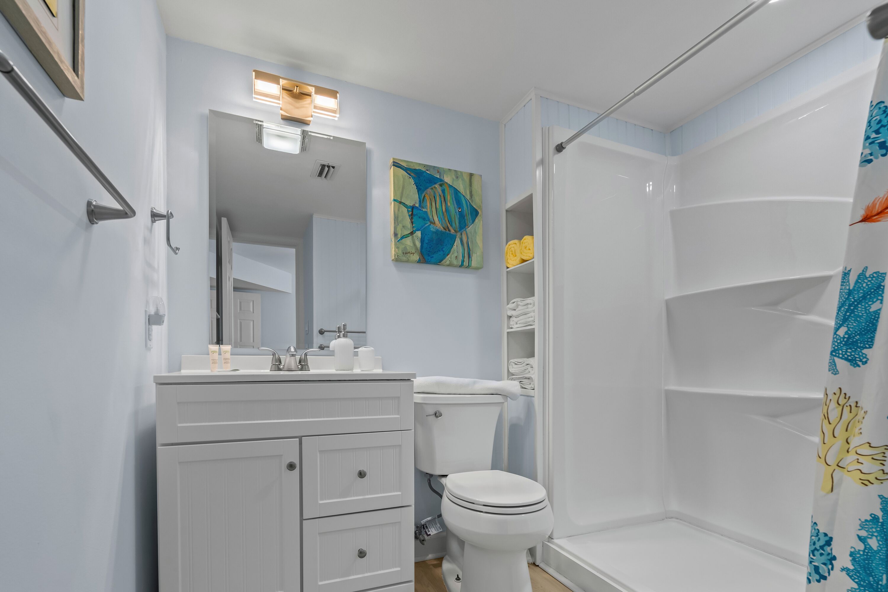 Une salle de bain avec un meuble-lavabo blanc, des toilettes, une douche avec un rideau de douche bleu et jaune, et un tableau accroché au mur.