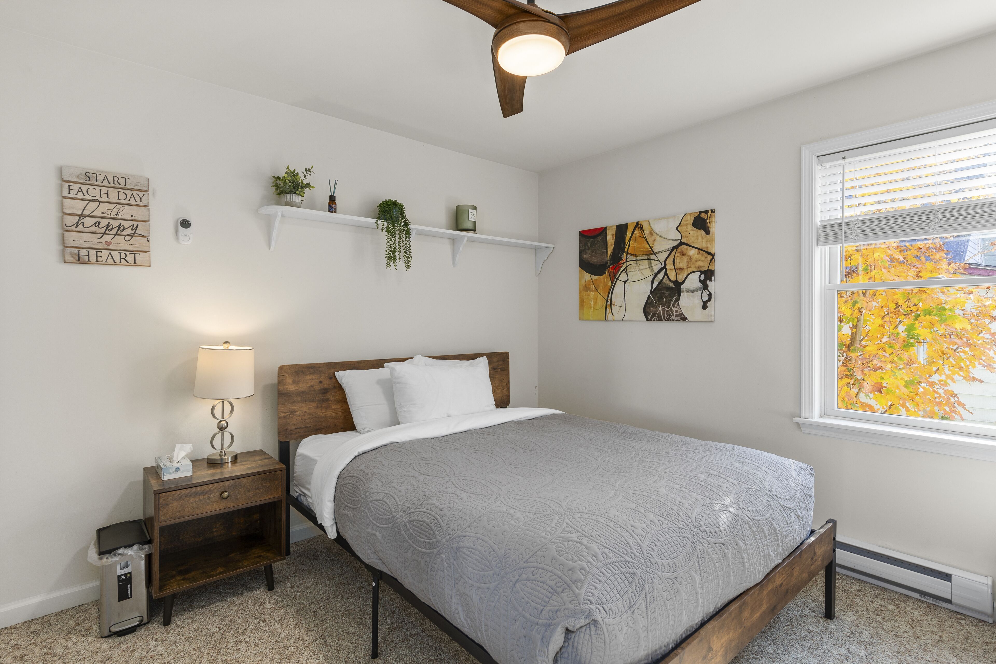 Une chambre à coucher comprenant un lit, une table de chevet, des éléments de décoration murale et une fenêtre donnant sur des arbres.