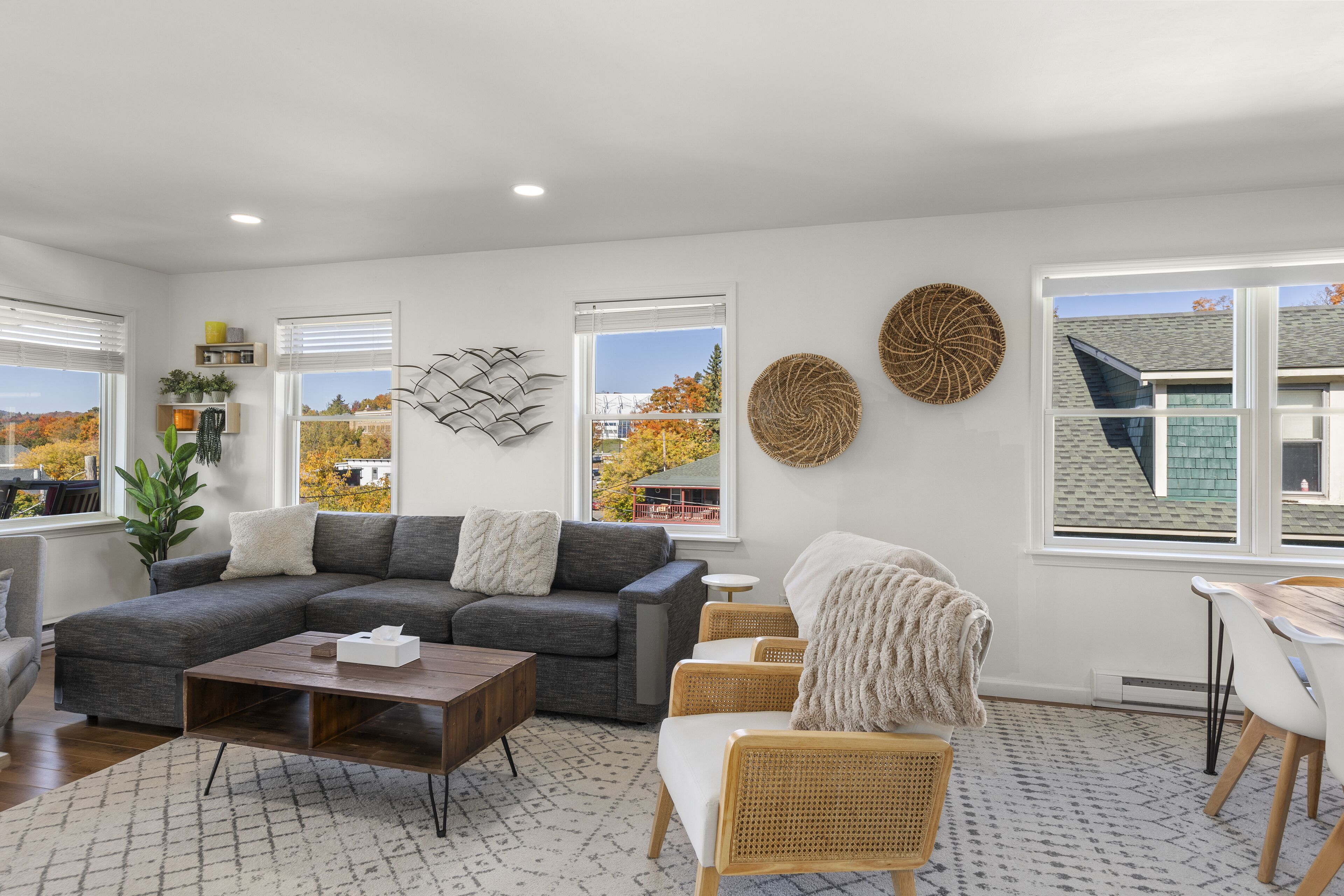 A modern living room with a sectional sofa, a coffee table, and a dining area with a table and chairs. There are large windows with blinds, decorative wall art, and a view of the outside.