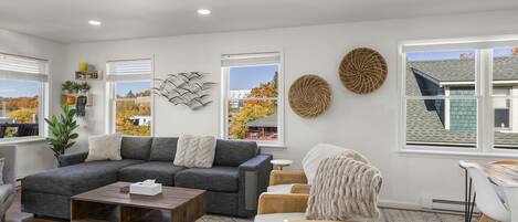 A modern living room with a sectional sofa, a coffee table, and a dining area with a table and chairs. There are large windows with blinds, decorative wall art, and a view of the outside.