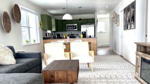 Una sala de estar moderna con sofá, sillón y mesa de centro. Se ve una cocina al fondo, con gabinetes y electrodomésticos verdes.