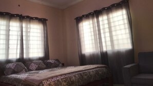 Un dormitorio con cama, dos ventanas con cortinas, un ventilador de techo y una silla.