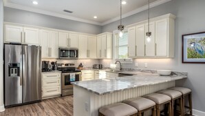 Una cocina moderna con armarios blancos, electrodomésticos de acero inoxidable, una isla de mármol con taburetes altos e iluminación colgante.
