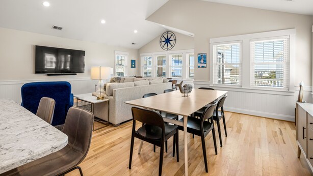 A modern living room with a dining area, a large window with blinds, a mounted TV, and a ceiling fan. - Close to beach, on golf course in TCC!  Private pool, spa, and 6 seat golf cart! (Corolla)