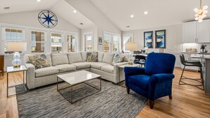 A spacious living room with a sectional sofa, a blue armchair, and a coffee table. There are large windows with blinds, a kitchen area, and a ceiling with recessed lighting. - Close to beach, on golf course in TCC!  Private pool, spa, and 6 seat golf cart! (Corolla)