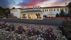 A hotel with a covered entrance, a well-maintained garden with pink flowers, and a clear sky.