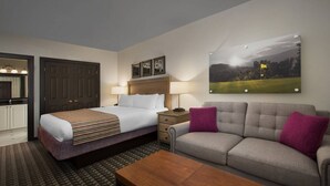 A hotel room with a bed, a sofa, a coffee table, a nightstand, a lamp, and a painting on the wall.