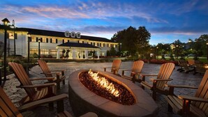 A patio area with a fire pit surrounded by Adirondack chairs, a covered seating area, and a building in the background.
