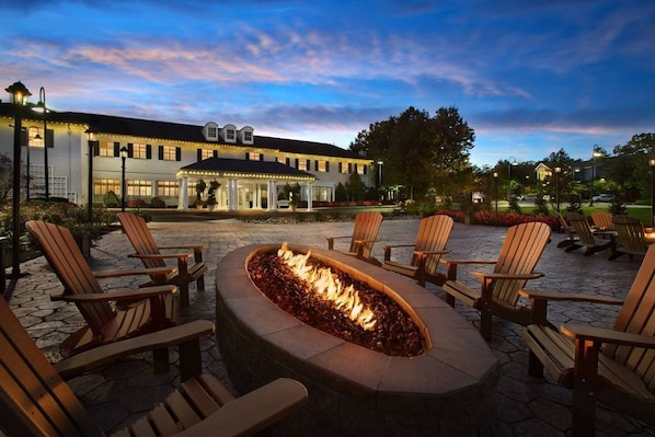 A patio area with a fire pit surrounded by Adirondack chairs, a covered seating area, and a building in the background.