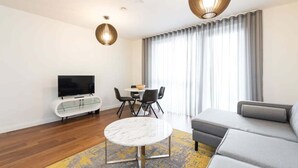 A modern living room with a sofa, a TV on a glass cabinet, a dining table with chairs, and a large window with curtains. - Fernbank by Dublin At Home (Dublin 14)