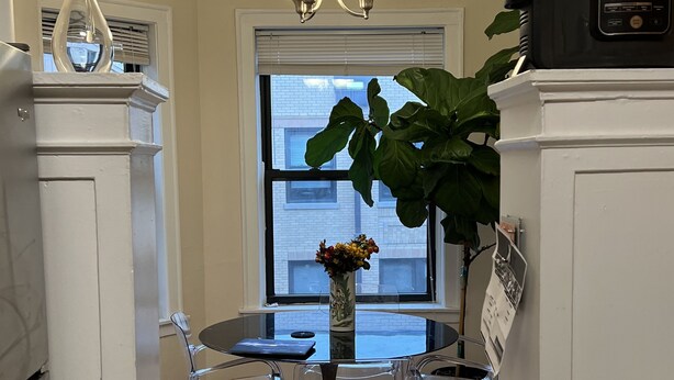 Una cocina con una mesa redonda de cristal, dos sillas transparentes, un jarrón con flores, un refrigerador y una cafetera.