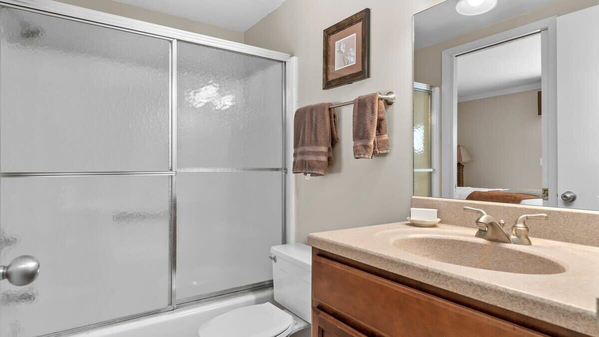 Un baño con cabina de ducha de vidrio, inodoro blanco, tocador con lavamanos y espejo, y dos toallas colgadas en la pared.