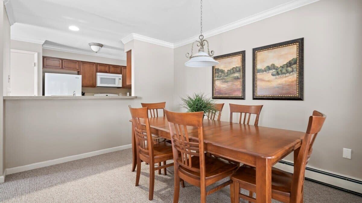 A dining area with a wooden table and chairs, a hanging light fixture, and two framed paintings on the wall.