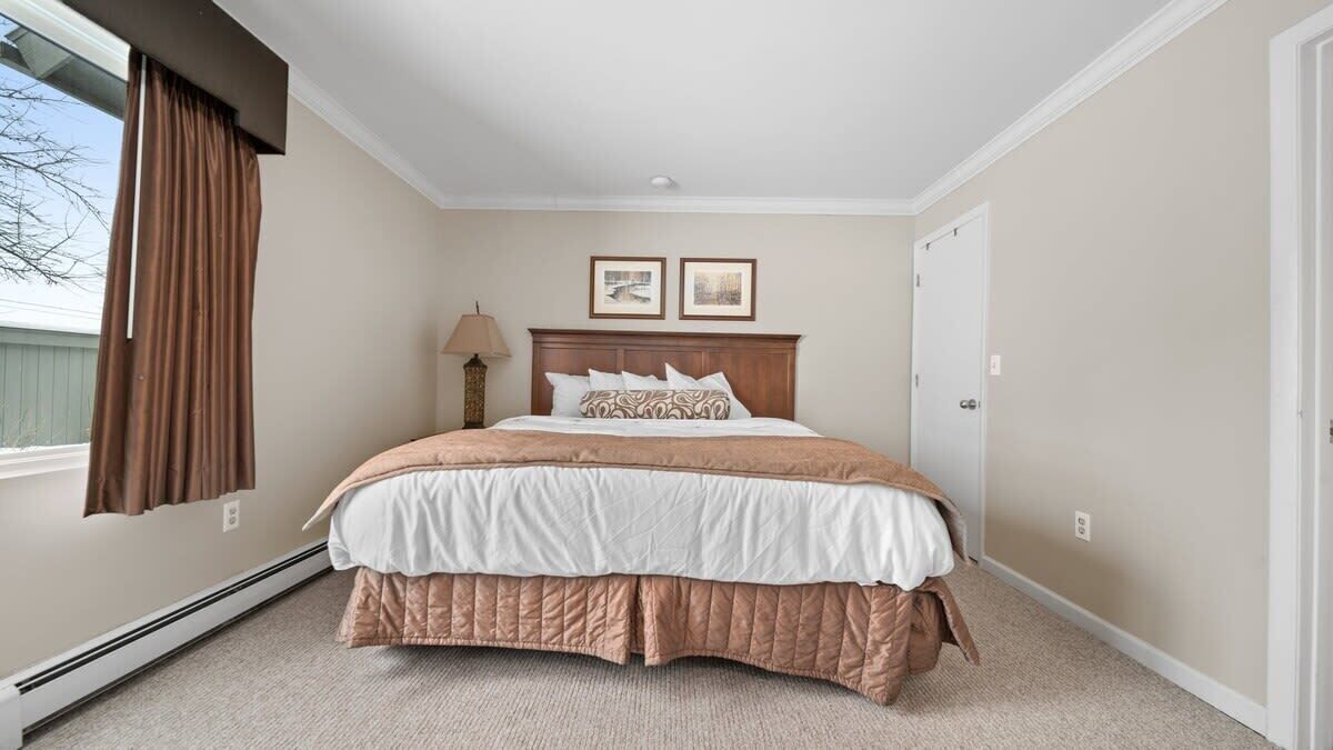 Un dormitorio con cama, ventana con cortinas, lámpara de pared y dos cuadros enmarcados.