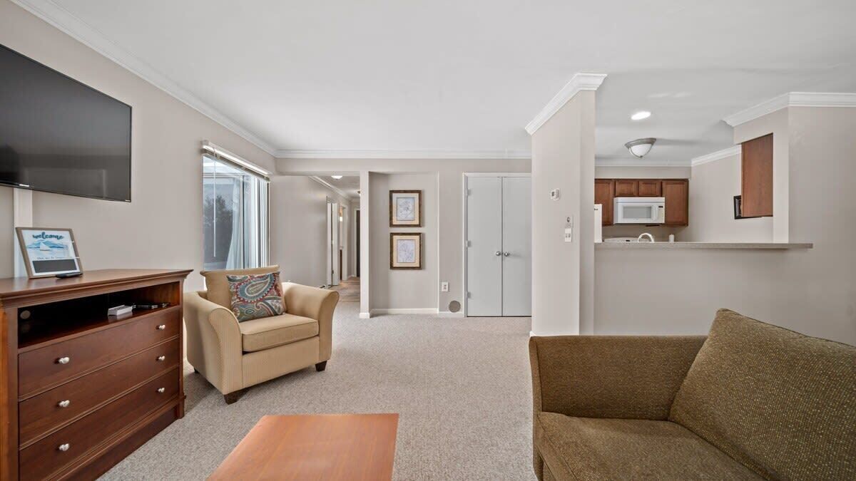 A spacious living room with a sofa, a coffee table, and a TV on the wall. There is a kitchen area with a microwave and cabinets.