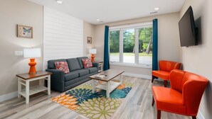 A modern living room with a sofa, a coffee table, and two armchairs. There is a TV mounted on the wall and a window with curtains. - Condo Near Downtown | King Bed + Dog Friendly (Traverse City)