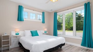 A bedroom with a large bed, two bedside tables, a window with curtains, and a view of the outdoors. - Condo Near Downtown | King Bed + Dog Friendly (Traverse City)
