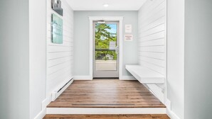A narrow hallway with white walls and wooden flooring leading to a door with a view of greenery outside. - 2BR Condo | King Bed + Shared Rooftop + Dogs (Traverse City)