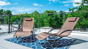 Two lounge chairs on a blue patterned deck with a glass balustrade and trees in the background. - 2BR Condo | King Bed + Shared Rooftop + Dogs (Traverse City)