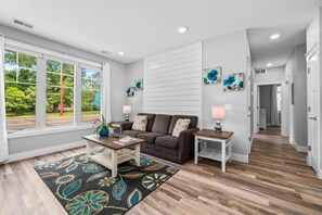 A modern living room with a sofa, coffee table, and side tables. There are large windows, a patterned rug, and artwork on the walls. - 2BR Condo | King Bed + Shared Rooftop + Dogs (Traverse City)