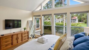 A bedroom with a large bed, a TV, a wooden dresser, and multiple windows offering views of a residential area. - **The Caddy's Shack** 1 story home with Club Cards Optional (Fripp Island)
