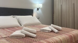 Una cama bien hecha con ropa blanca y toallas enrolladas, un cabecero y una lámpara fijada a la pared.