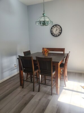A dining area with a wooden table and chairs, a wall clock, and a hanging light fixture.