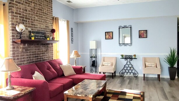 A living room with a red sofa, a coffee table, a patterned rug, a ceiling fan, and a brick wall. - Cozy 3 bedroom house (Indianapolis)