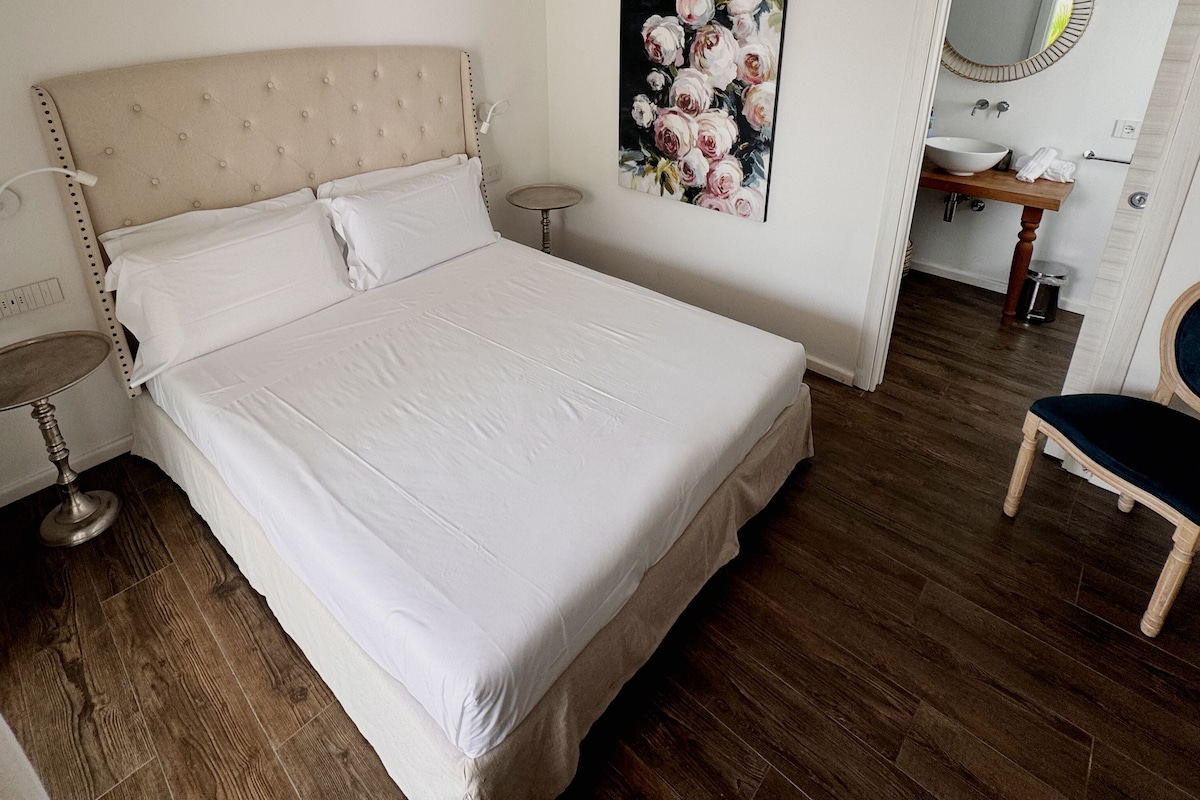A neatly made bed with a headboard, a small round table with a lamp, and a chair with a cushion.