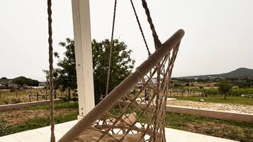 A rope swing chair with a netted seat, attached to a white pillar, overlooking a vineyard and distant hills.