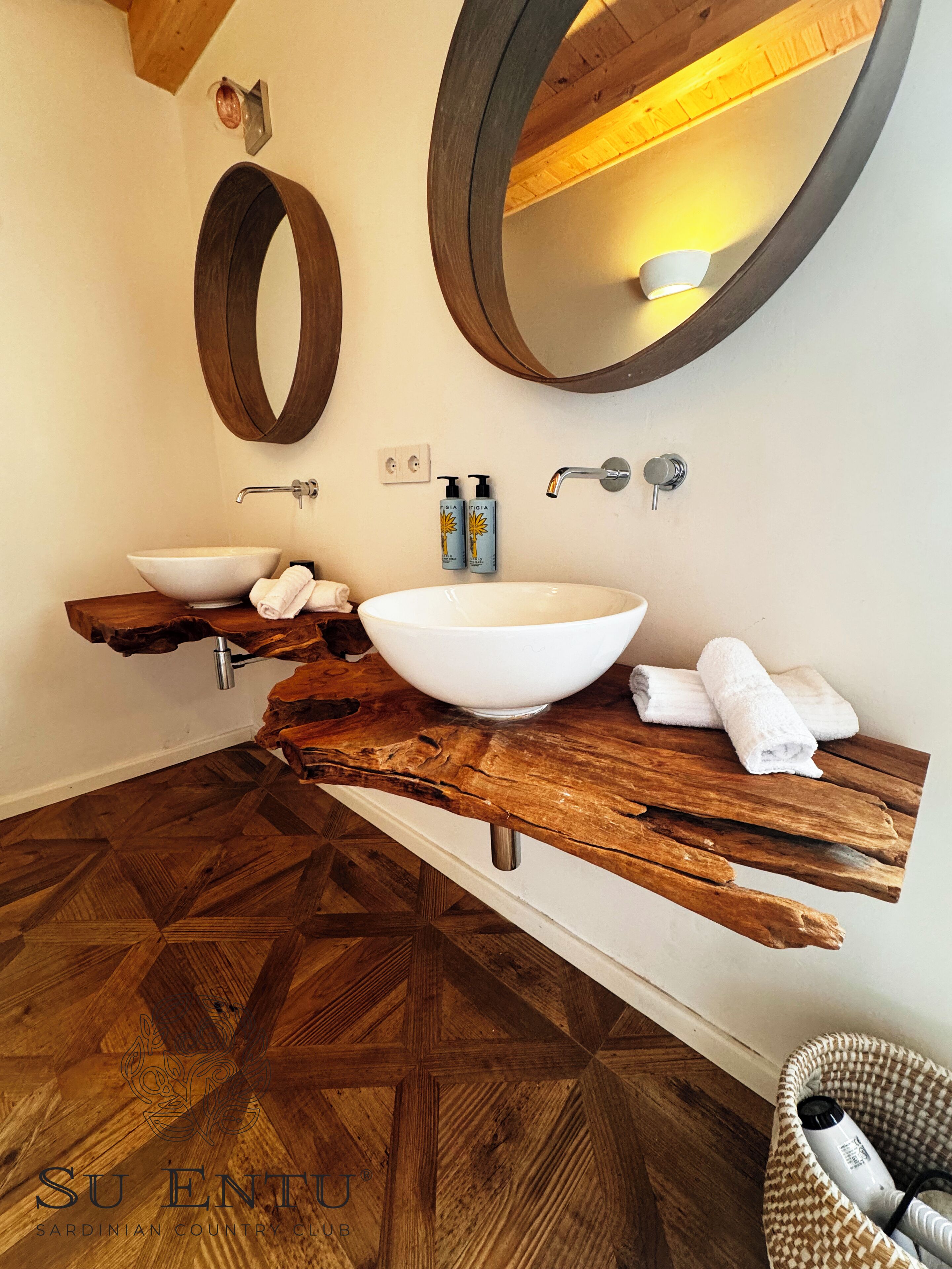 A bathroom with a wooden vanity, two large oval mirrors, a white vessel sink, and a wooden floor with a geometric pattern.
