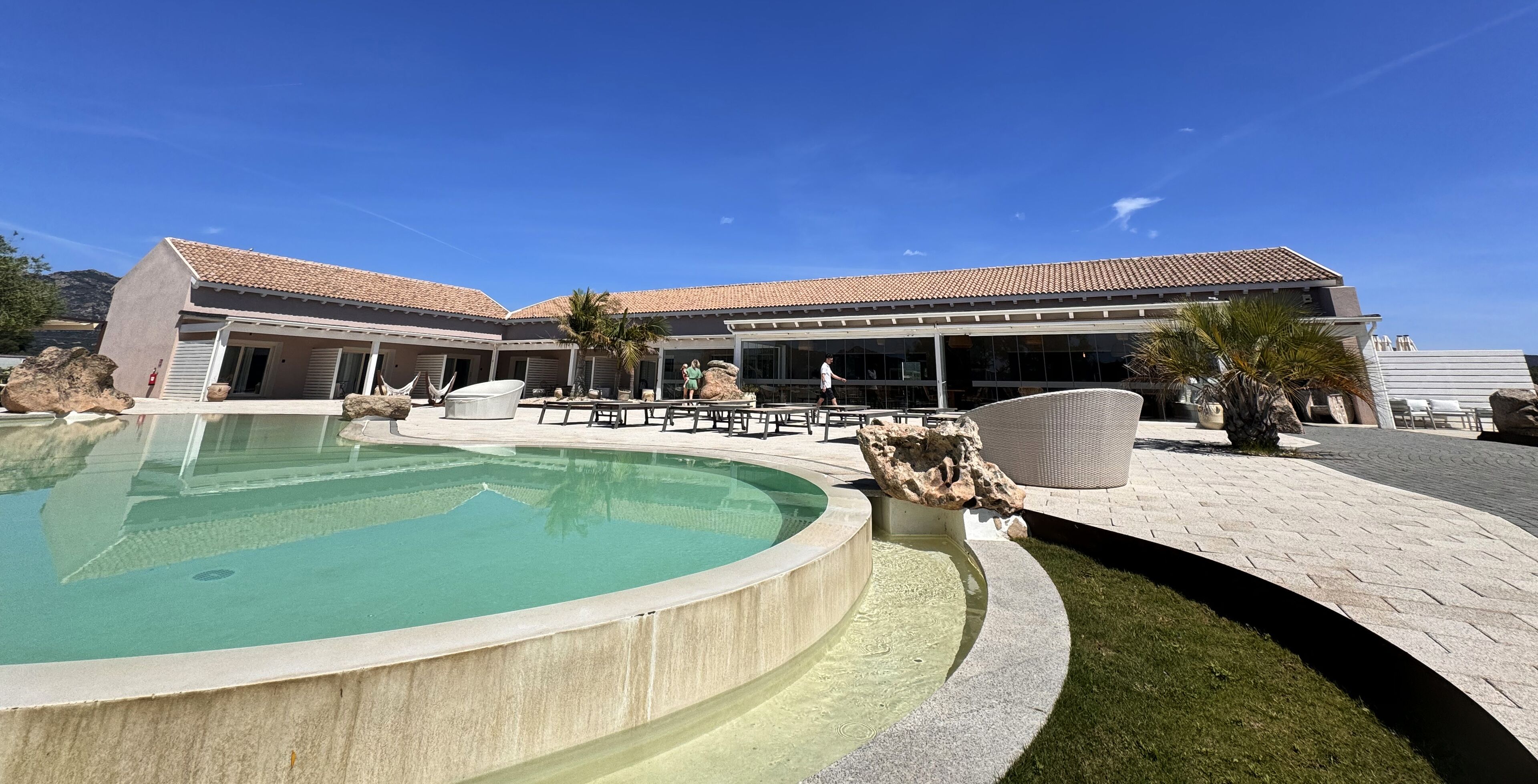 Ein Poolbereich mit einem runden Pool, Liegestühlen und einem Gebäude mit einem Ziegeldach.