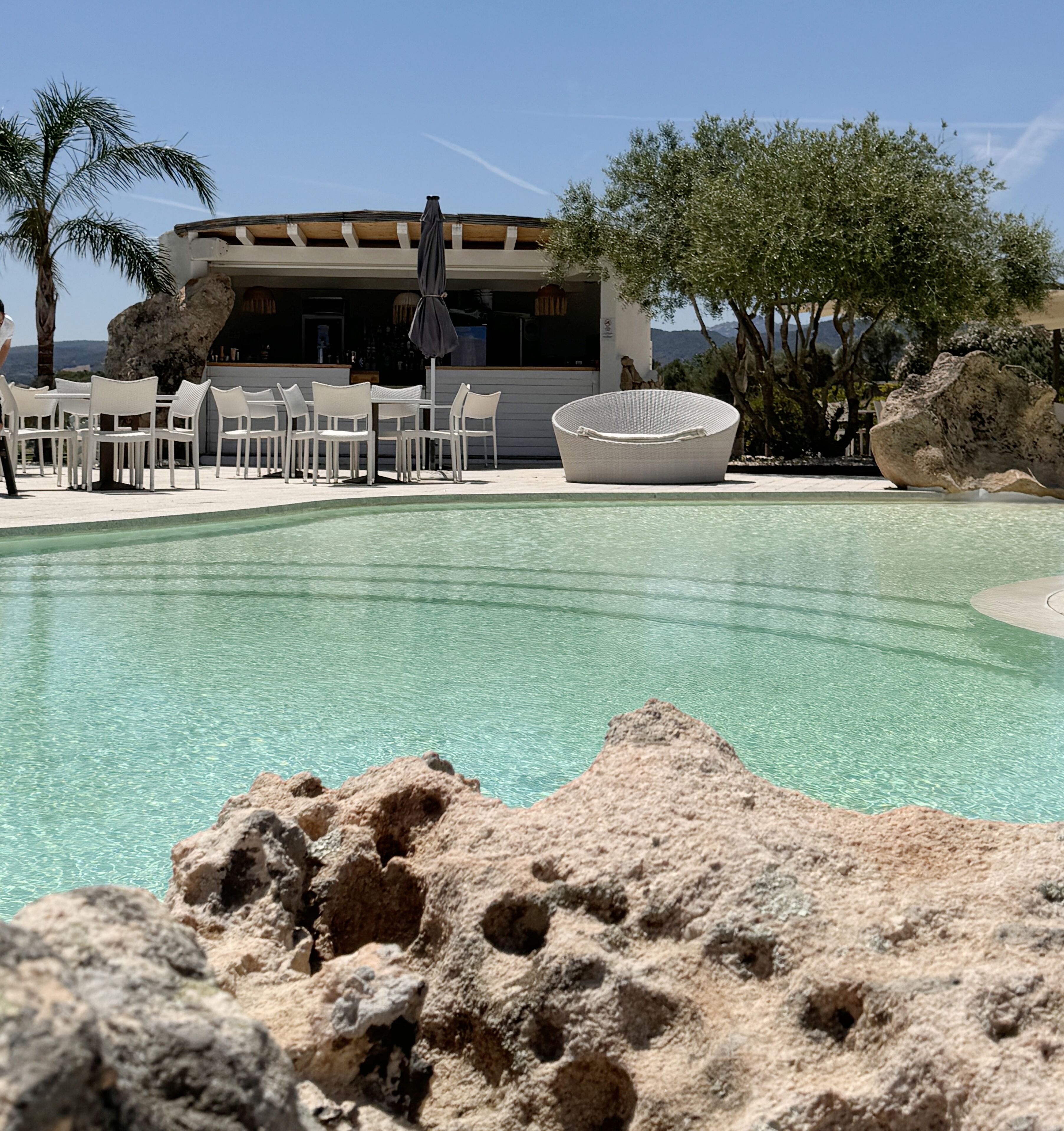 Una piscina con agua cristalina, rodeada de rocas y mobiliario exterior blanco.