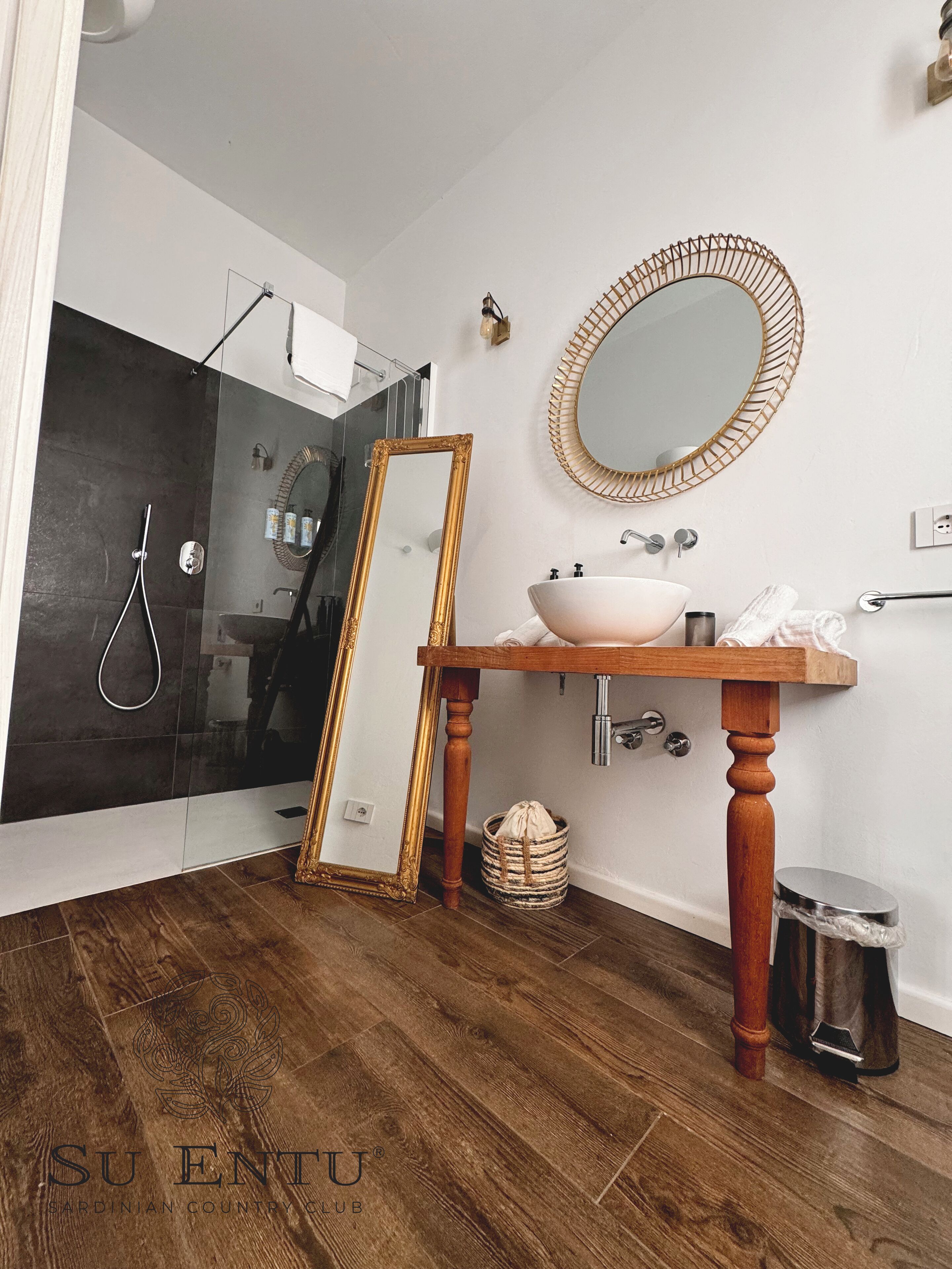 Un baño con tocador de madera, un espejo redondo, cabina de ducha de vidrio y zona de ducha con azulejos oscuros.