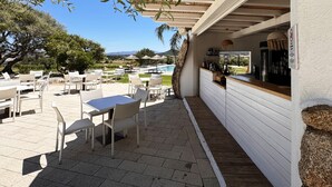 Un patio exterior con mobiliario de mimbre blanco, una piscina y una zona de bar con botellas y un refrigerador.
