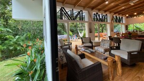 A spacious indoor area with wooden flooring, multiple seating arrangements, and large windows offering a view of the surrounding greenery. - SAMARA - Stunning 2 bedroom House 15 min from the Beach. (Sámara)