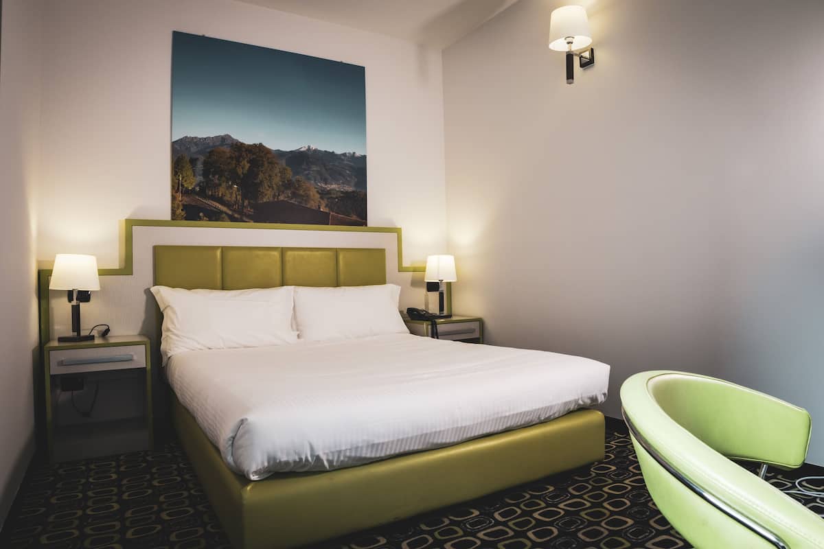 A hotel room with a bed, two bedside lamps, a green chair, and a painting on the wall.