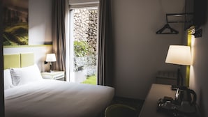 Hotellihuone, jossa on sänky, yöpöytä lampun kanssa, työpöytä lampun kanssa, naulakko ja ikkunasta avautuva näkymä puutarhaan.