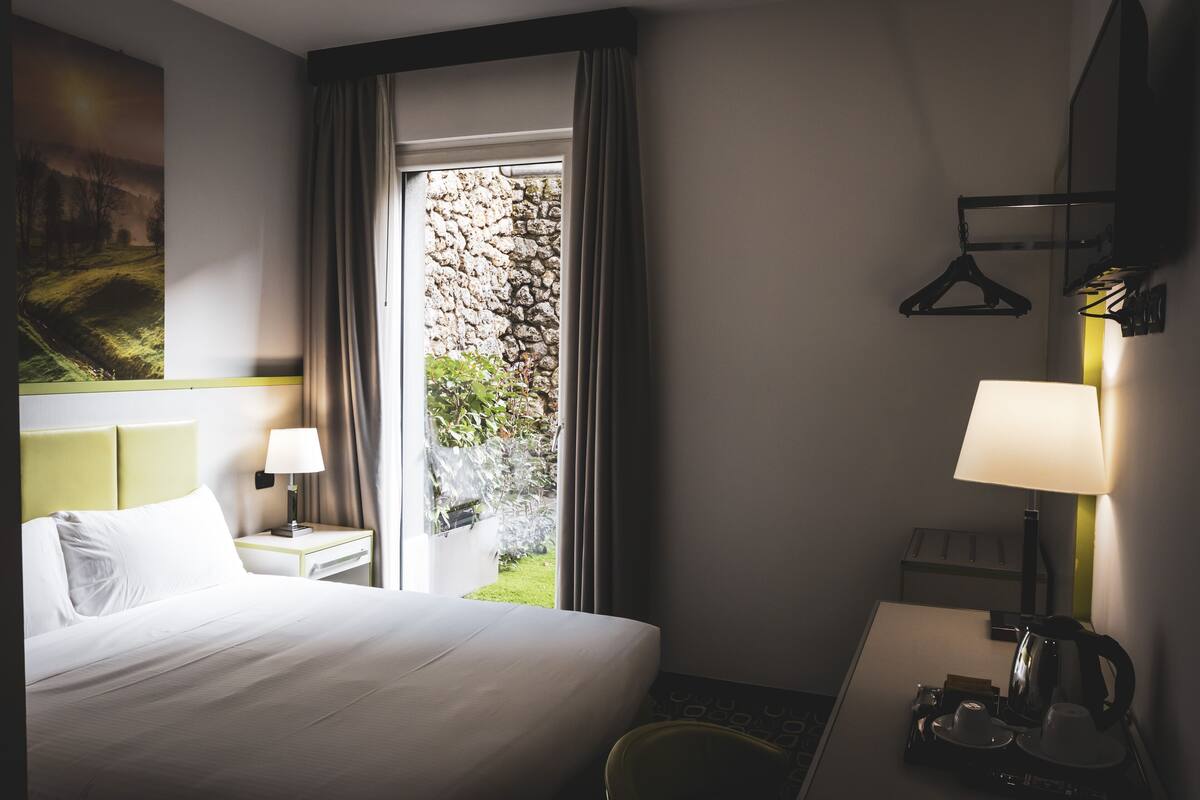 A hotel room with a bed, a nightstand with a lamp, a desk with a lamp, a coat rack, and a view of a garden through a window.