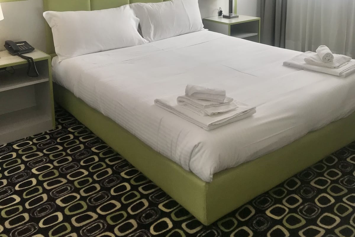 A hotel room with a neatly made bed, a painting of a natural scene, a telephone, and a nightstand with a lamp.