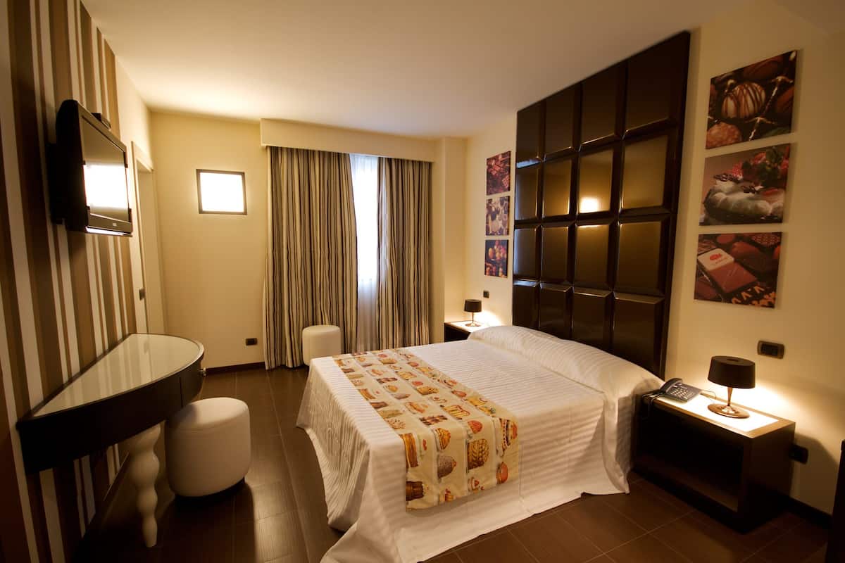 A hotel room with a bed, a desk, a chair, a TV, a nightstand with a lamp, and a wall with framed pictures.
