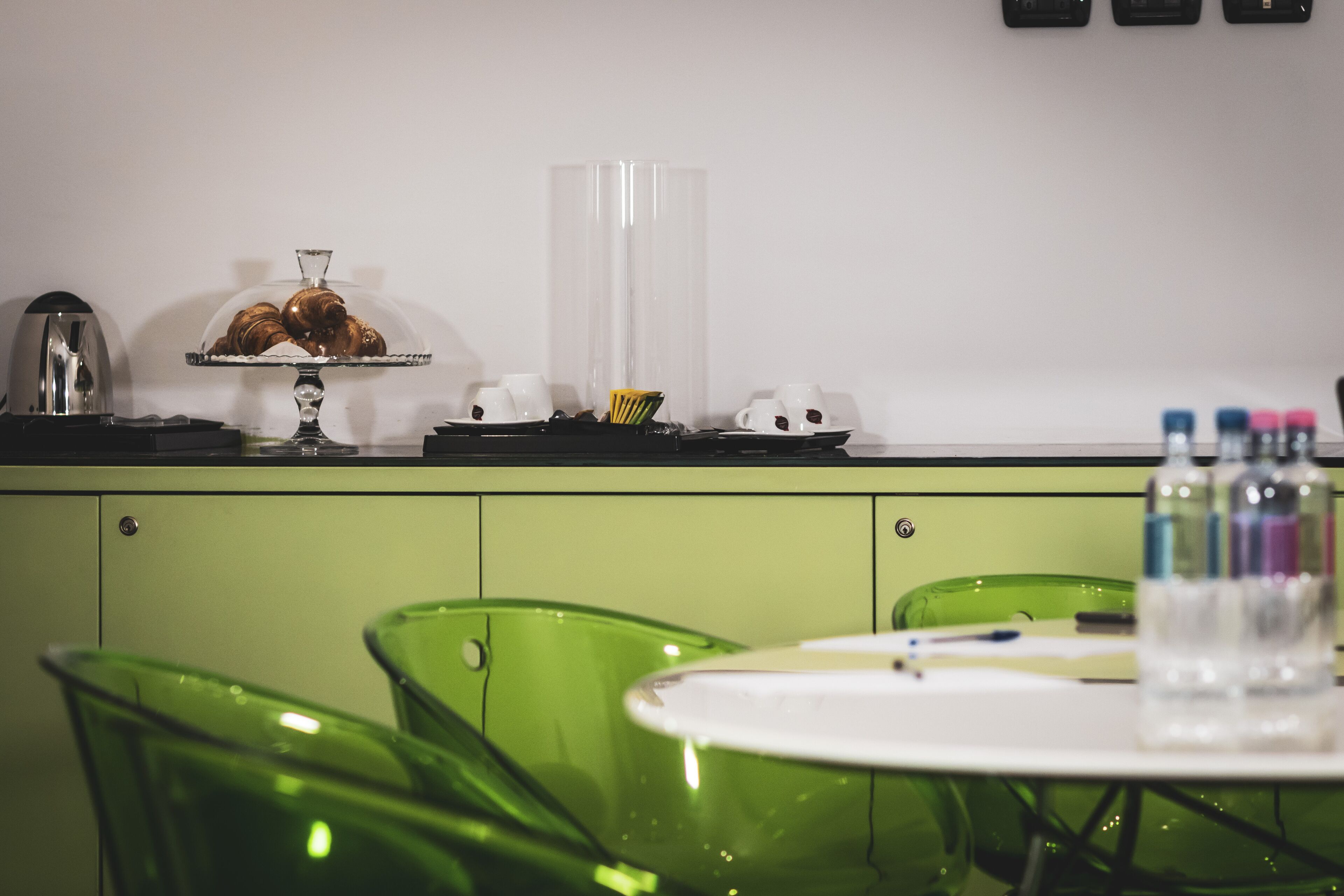 A green dining area with a glass display case containing pastries, a coffee station, and a water bottle on the table.