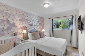 Ein Schlafzimmer mit einem Bett, einem Babybett, einer Wand mit Blumenmuster, einem Fenster mit Vorhängen und einem an der Wand befestigten Fernseher.