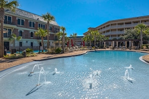 A swimming pool with water fountains, surrounded by palm trees and a multi-story building with balconies.
